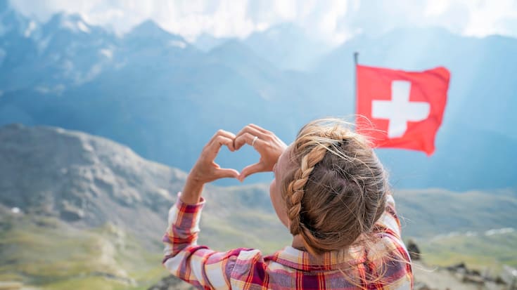 Young woman making heart shape finger frame on mountain top overlooking the Swiss Alps; Swiss flag