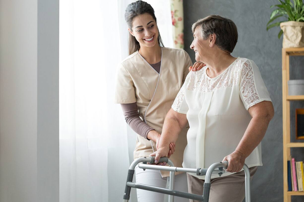 Smiling beautiful caregiver and disabled old lady with walker at home